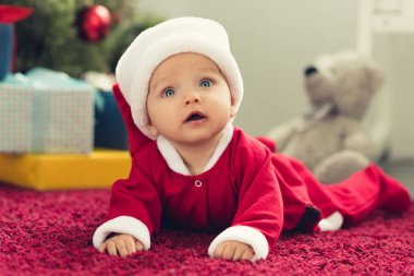 adorable little baby in santa suit lying on red carpet with christmas gifts blurred on background and looking up clipart
