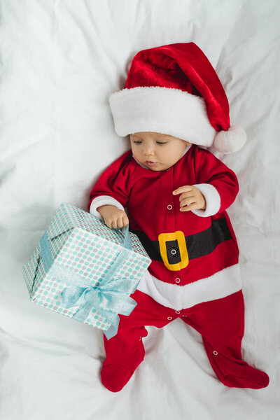 top view of adorable little baby in santa suit lying in crib with christmas gift