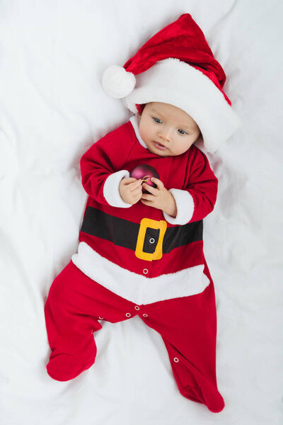 top view of adorable little baby in santa suit holding christmas bauble while lying in crib