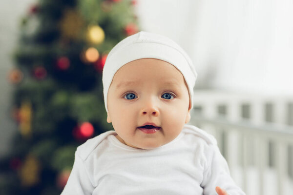 close-up portrait of adorable little baby in white hat looking at camera with blurred christmas tree on background