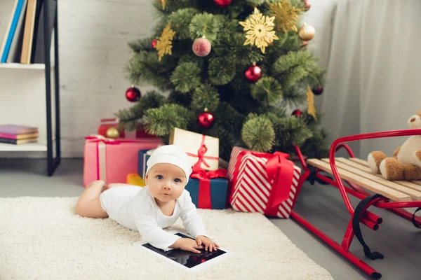 可爱的小婴儿与片剂躺在地板附近圣诞礼物和树 看着相机 — 图库照片