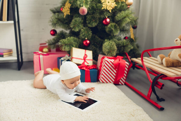 adorable little baby with tablet lying on floor near christmas gifts and tree
