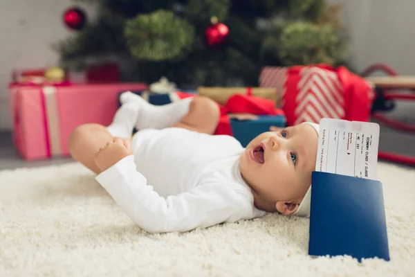 航空券とパスポートの背景にぼやけているクリスマス ツリーを床に横たわって幸せな赤ちゃん — ストック写真