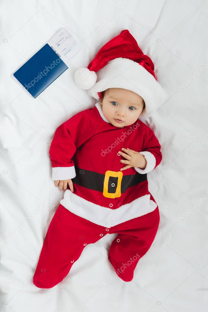 top view of adorable little baby in santa suit lying in crib with flight ticket and passport