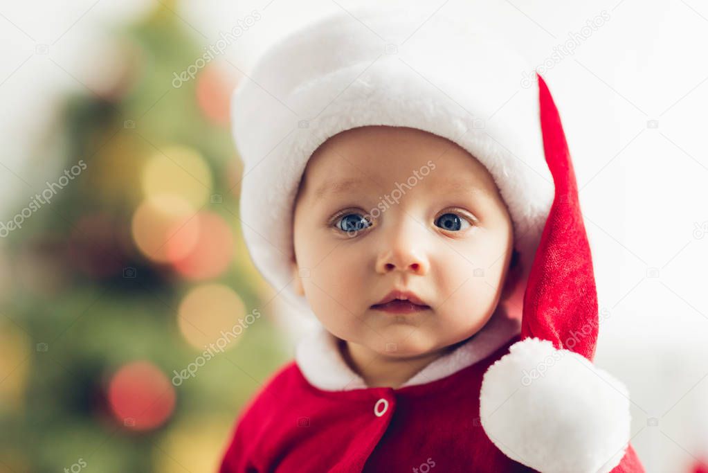 close-up portrait of cute little baby in santa hat looking at camera with blurred christmas tree on background