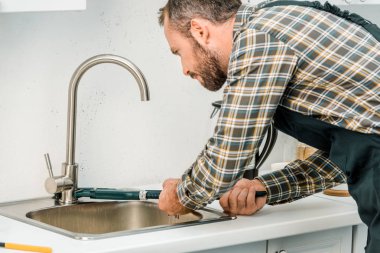 handsome plumber repairing tap with monkey wrench in kitchen clipart
