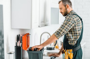 side view of handsome plumber taking tools from toolbox in kitchen clipart