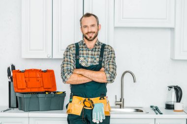 çapraz kolları ile ayakta ve mutfak kamerada bakarak gülümseyen yakışıklı tesisatçı