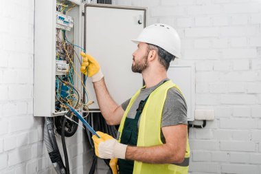 side view of electrician holding clipboard and checking wires in electrical box in corridor clipart