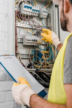 cropped image of electrician holding clipboard and inspecting electrical box in corridor clipart