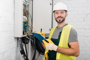 smiling handsome electrician holding clipboard near electrical box in corridor and looking at camera clipart