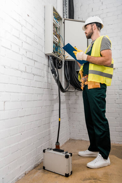 low angle view of handsome electrician holding clipboard and checking electrical box in corridor