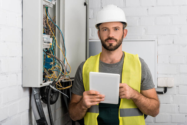 handsome electrician holding tablet near electrical box in corridor and looking at camera