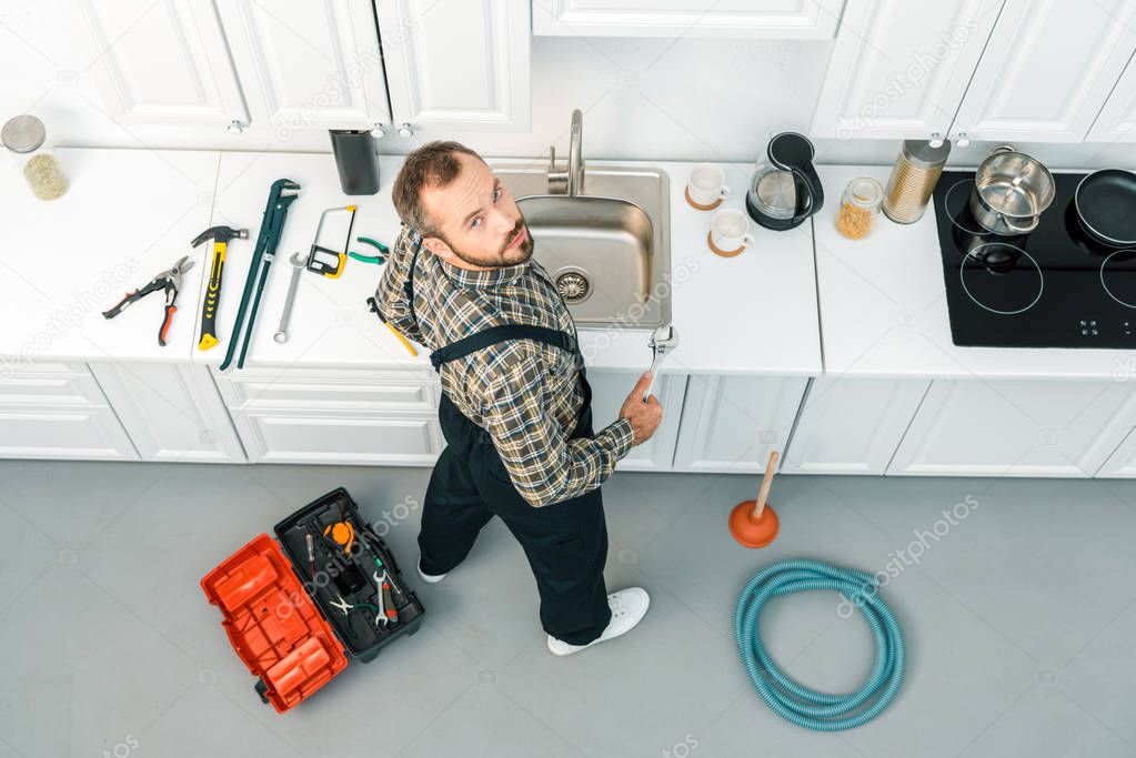 high angle view of handsome plumber holding adjustable wrench and looking at camera in kitchen