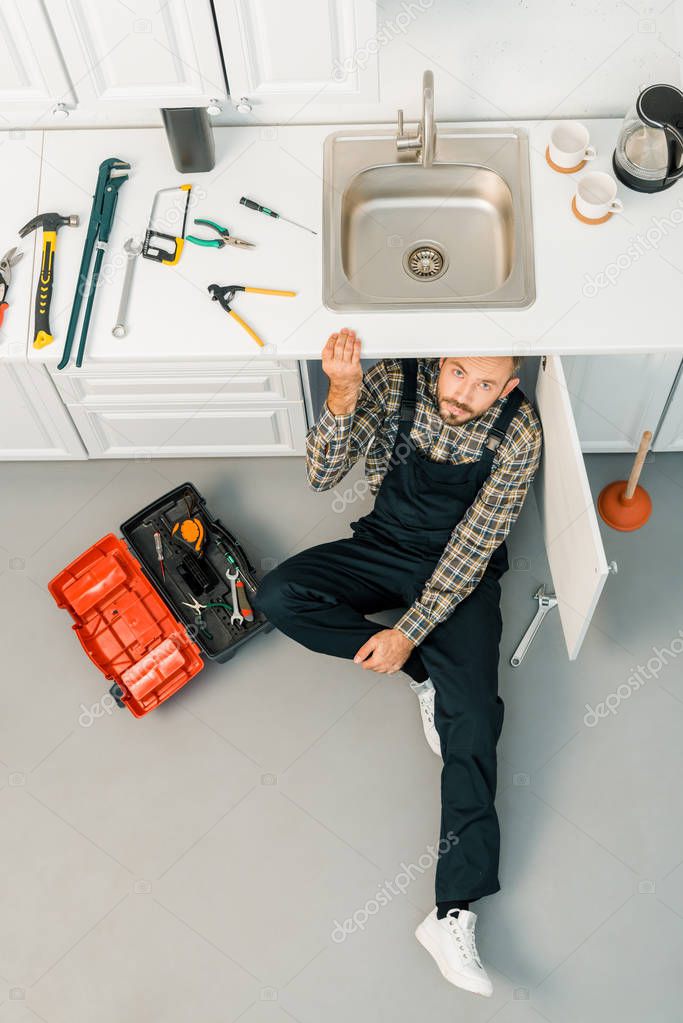 high angle view of handsome plumber sitting on floor near sink and looking at camera in kitchen