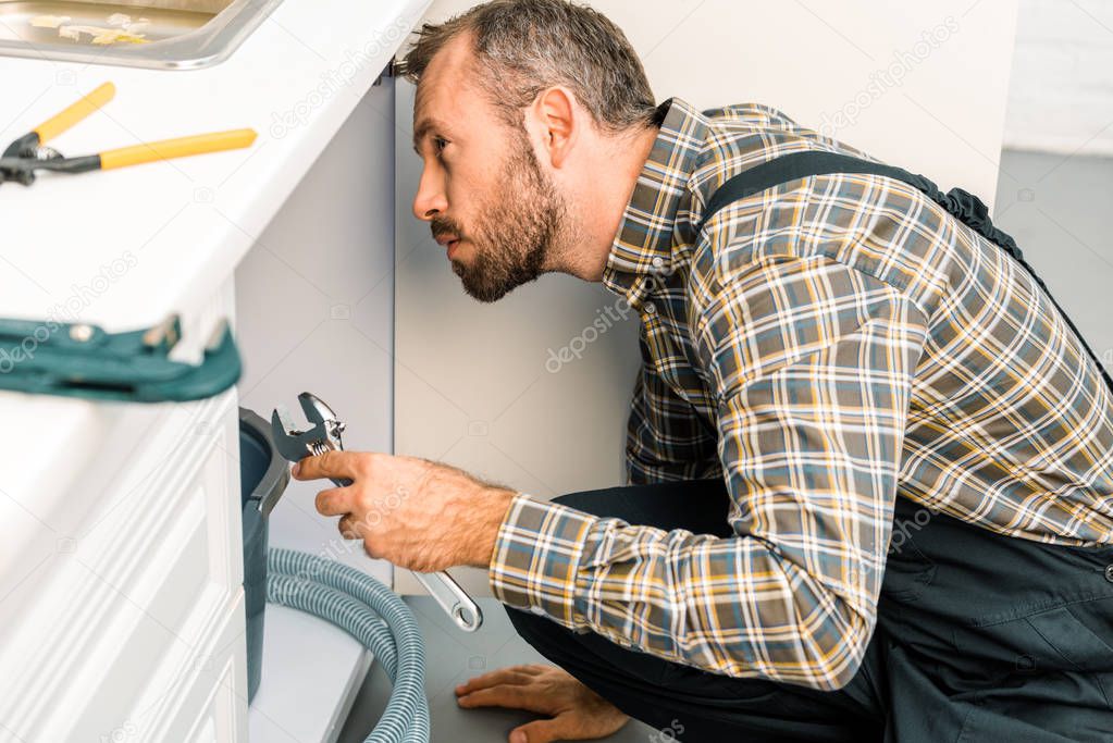 side view of handsome plumber holding adjustable wrench and looking under broken sink in kitchen