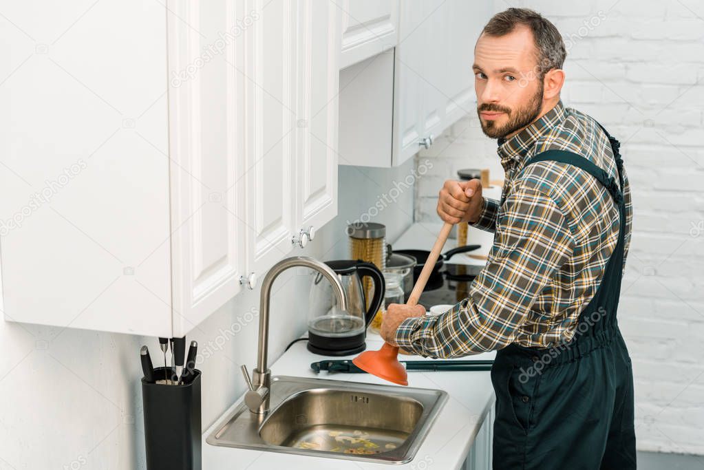 handsome plumber using plunger and cleaning sink in kitchen, looking at camera