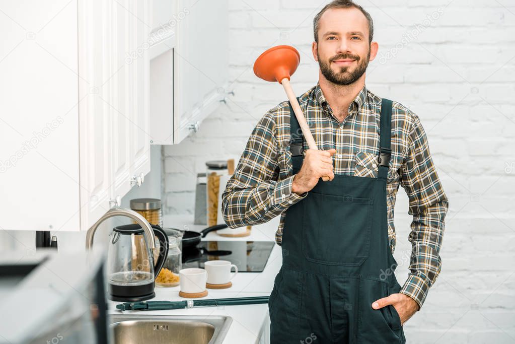 cheerful handsome plumber holding plunger on shoulder and looking at camera in kitchen