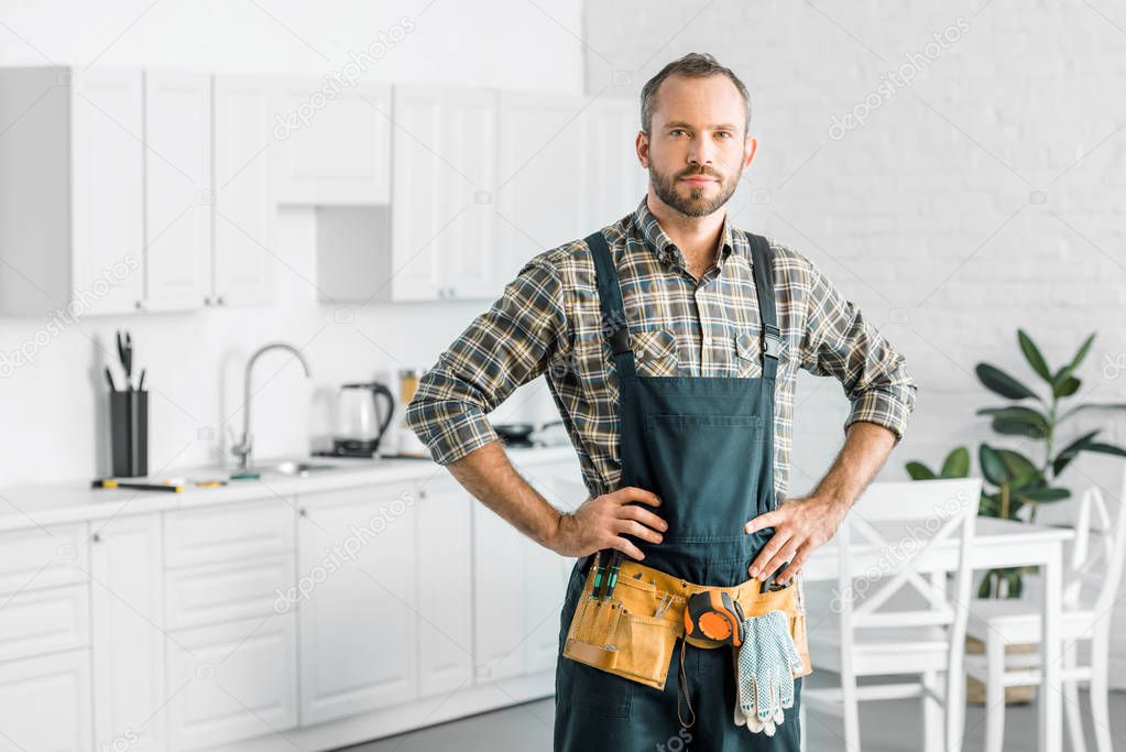 handsome plumber in overalls and tool belt looking at camera in kitchen