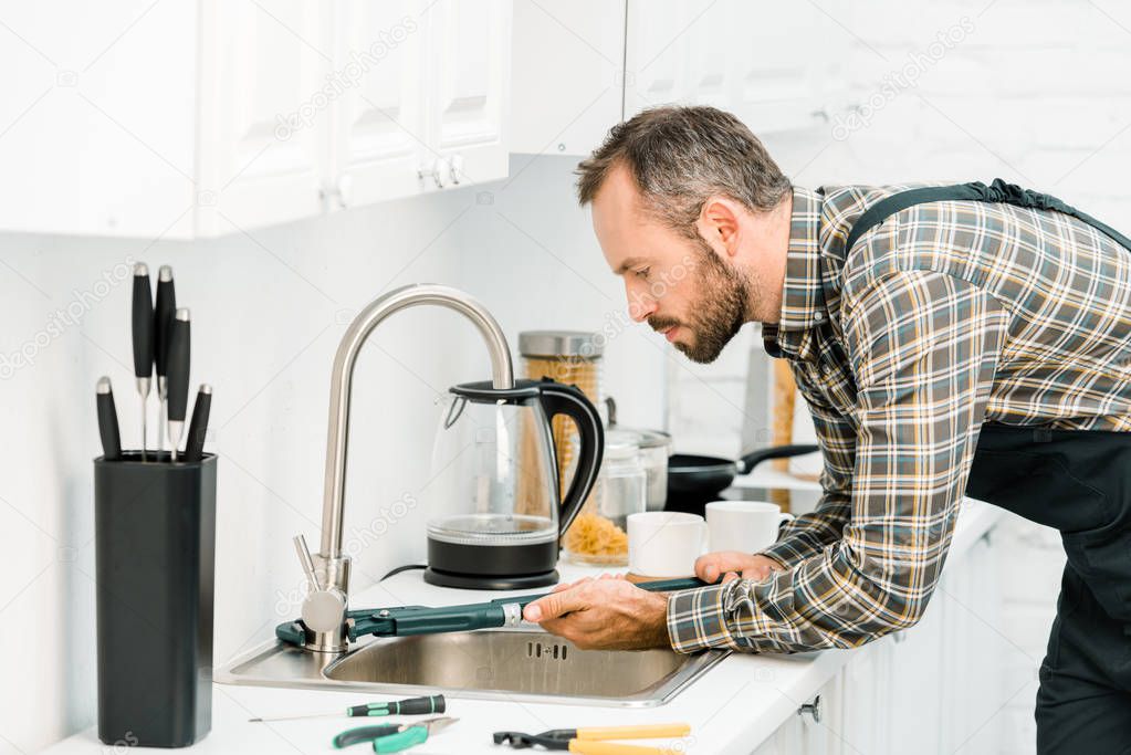 side view of handsome plumber repairing tap with monkey wrench in kitchen