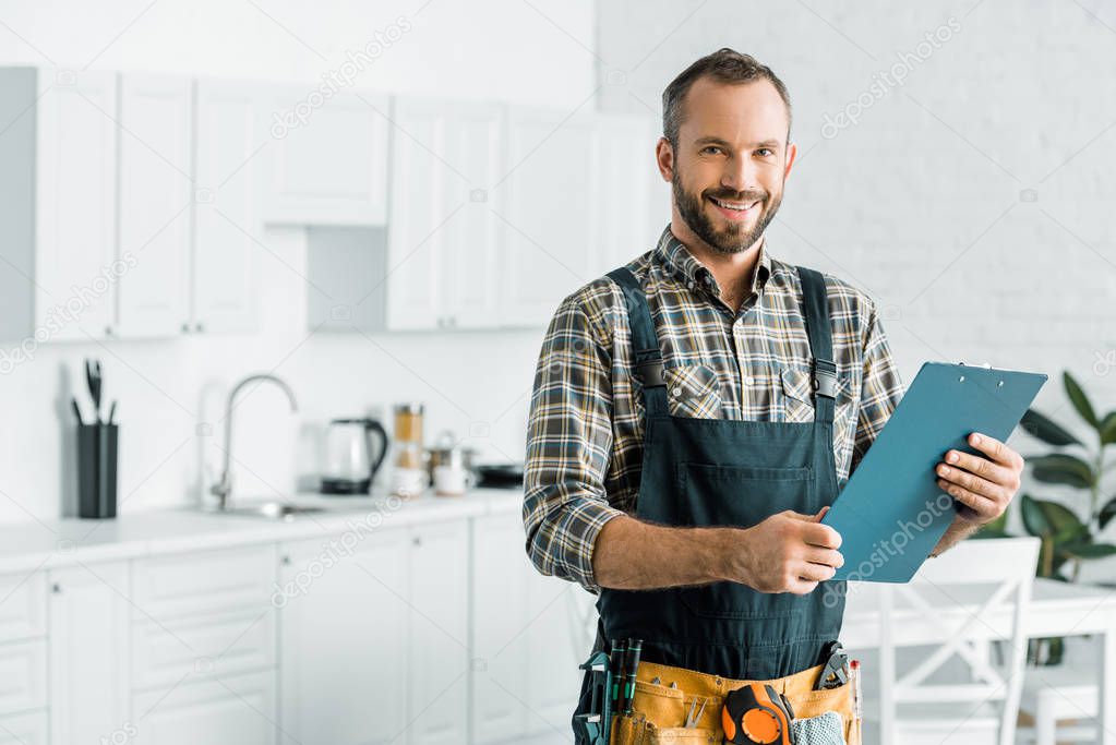 smiling handsome plumber holding clipboard and looking at camera in kitchen