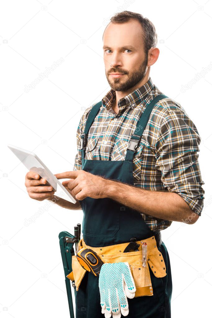 handsome plumber with tool belt using digital tablet isolated on white and looking at camera