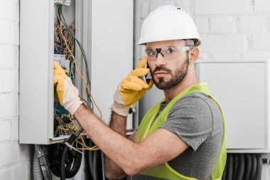 surprised electrician checking electrical box and talking by smartphone in corridor clipart