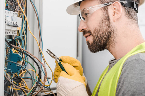 side view of handsome electrician holding pliers and looking at electrical box in corridor