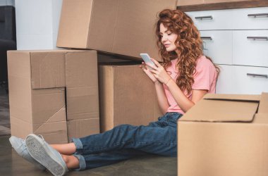 attractive woman with curly hair sitting on floor near cardboard boxes and using smartphone at new home clipart