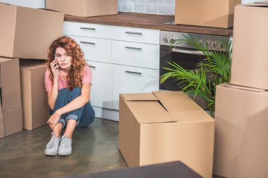 sad woman in casual clothes sitting on floor near cardboard boxes and talking by smartphone at new home clipart