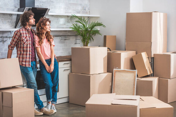 girlfriend and boyfriend with curly hair standing near cardboard boxes and looking away at new kitchen