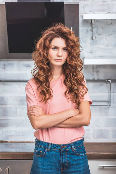 beautiful woman with red curly hair standing with crossed arms near cardboard boxes at new kitchen and looking at camera