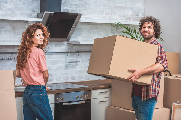 smiling couple unpacking cardboard boxes together at new kitchen and looking at camera