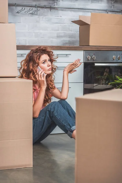 愤怒的女人 卷曲的头发坐在地板附近的纸板盒和智能手机交谈在新的厨房 — 图库照片