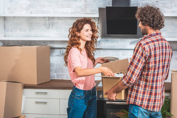 smiling couple unpacking utensil from cardboard box at new kitchen and looking at each other