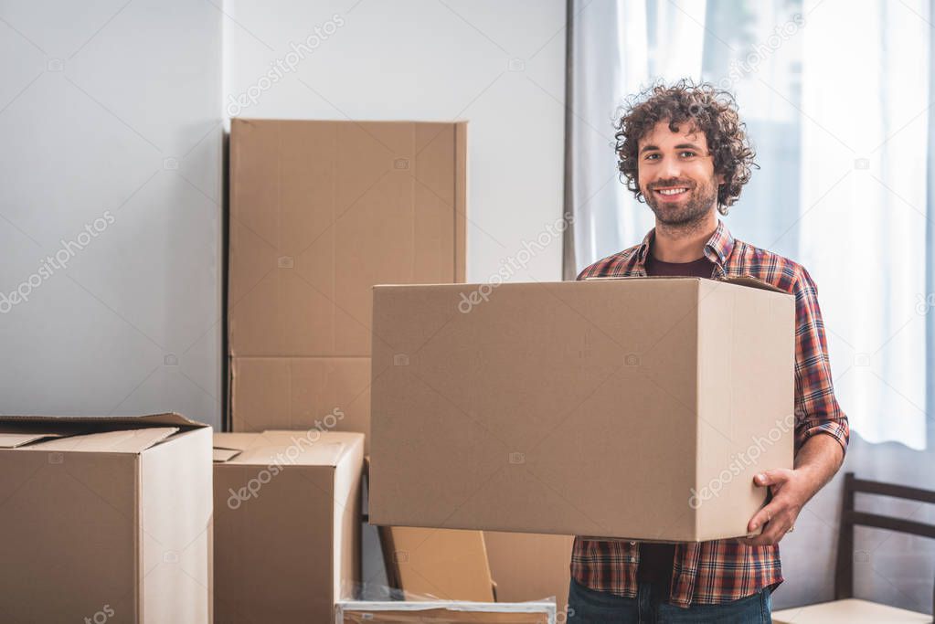 smiling handsome man with curly hair holding cardboard box at new home