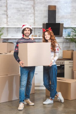 karton kutu tutarak ve kamera Noel, yeni evinde dururken gülümseyen mutlu genç çift