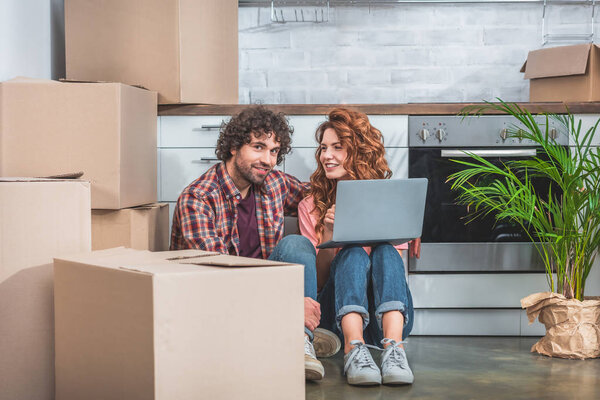 smiling couple sitting with laptop near cardboard boxes on floor in new kitchen