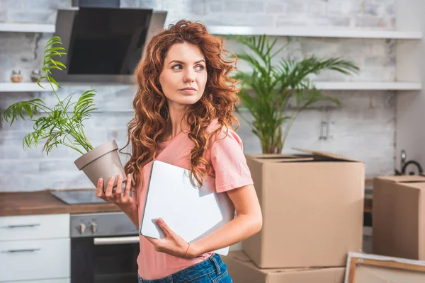 新しい家で鉢植えの植物とラップトップを保持している赤い巻き毛の美しい女性 — ストック写真