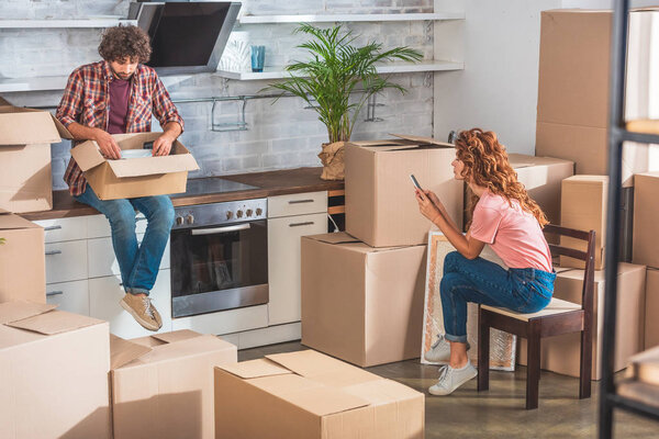 boyfriend unpacking utensil from cardboard box at new home, girlfriend using smartphone