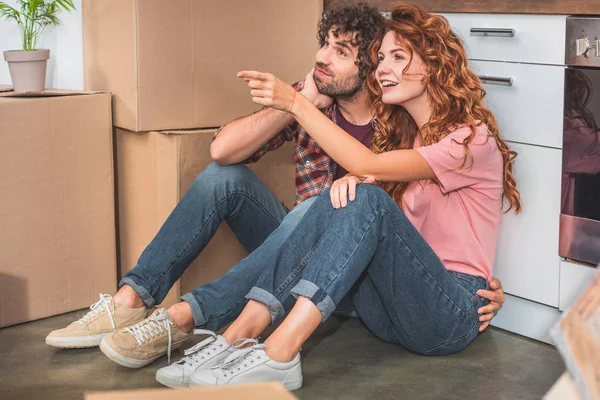 新しい台所の何かを指しているガール フレンドの段ボール箱の近くの床の上に座ってカップル — ストック写真