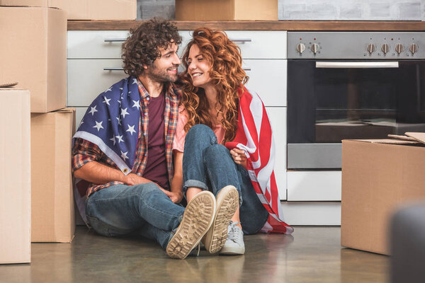 happy couple sitting wrapped in american flag near cardboard boxes at new home and looking at each other