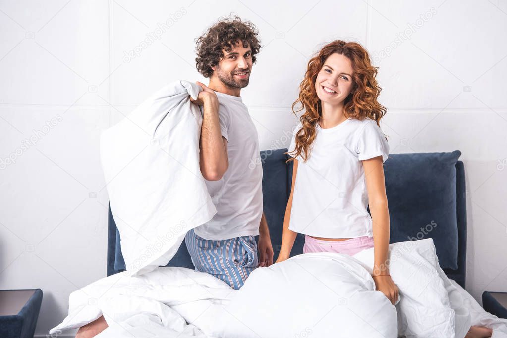 happy young couple holding pillows and smiling at camera in bedroom