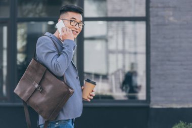 yakışıklı Asya adam deri sırt çantası ve kahve ile smartphone üzerinde konuşmaya başladın 