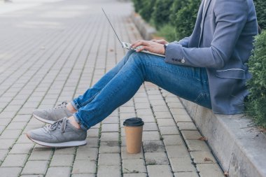 cropped view of freelancer with coffee to go working on laptop while sitting on sidewalk clipart