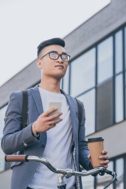 Kağıt bardak kahve ve smartphone Bisiklet yakınındaki şehirde tutarak kendine güvenen Asyalı adam