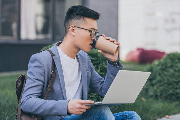handsome asian freelancer drinking coffee to go and working on laptop while sitting on sidewalk