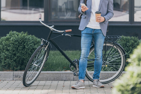 cropped view of stylish man with coffee to go using smartphone and standing near bike