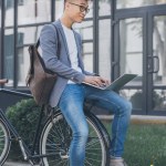 Pensive asian teleworker with leather backpack using laptop while sitting on bicycle