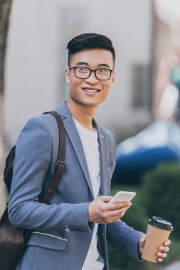 Tek kullanımlık bardak kahve tutarak ve smartphone kullanarak Asya adam gülümseyen 